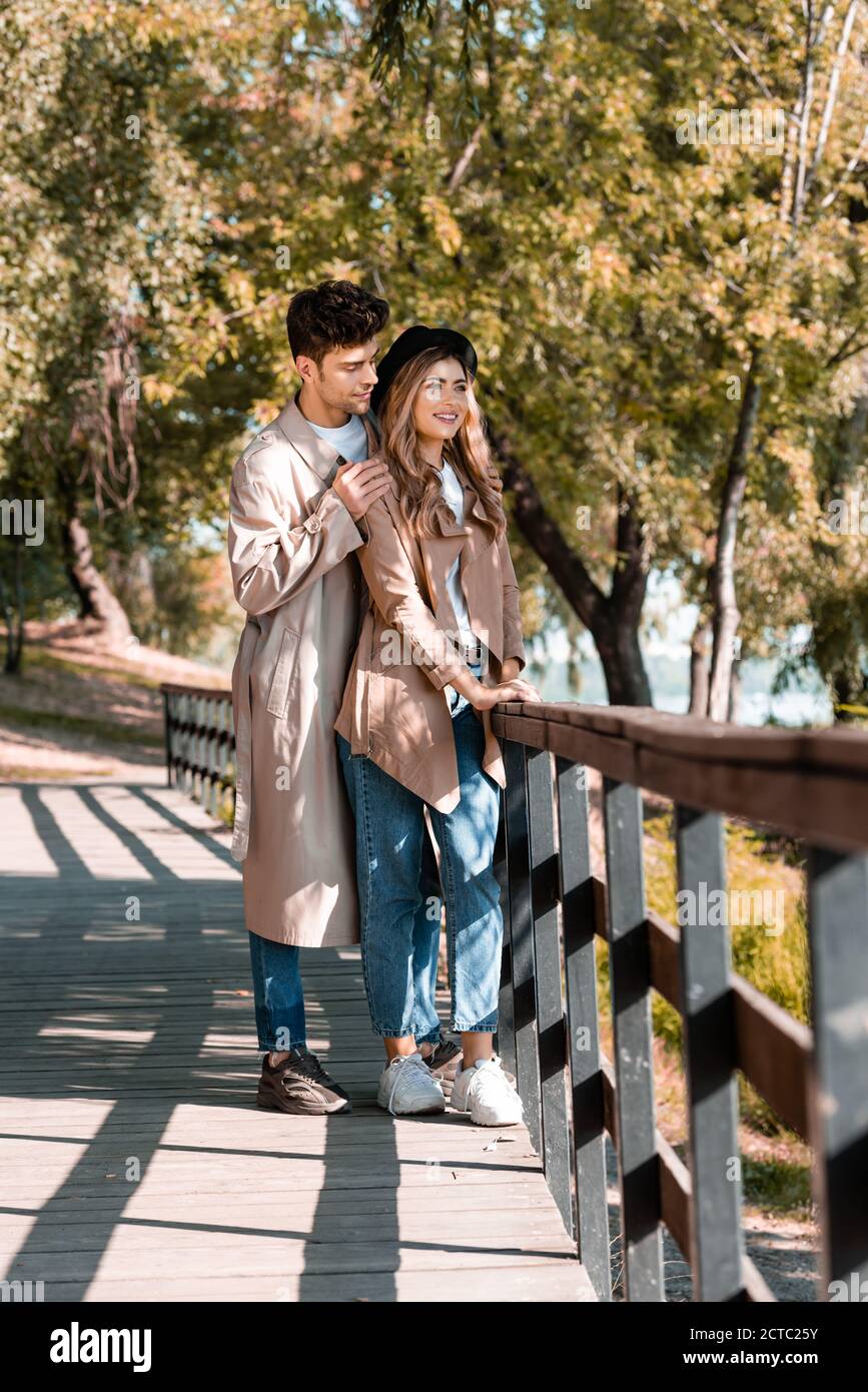 man touching shoulders of woman in hat and trench coat standing on wooden bridge in autumnal park Stock Photo