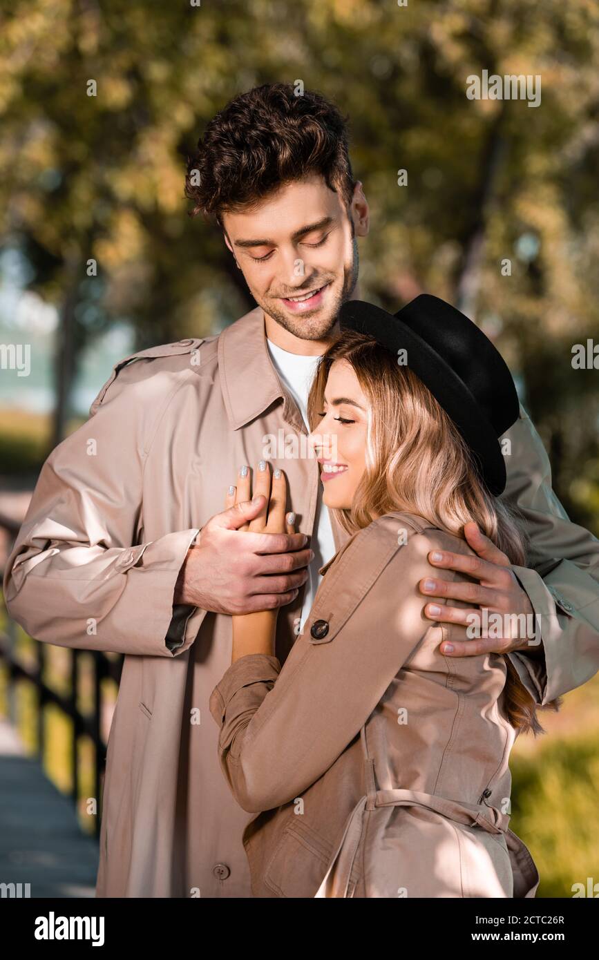 man in trench coat hugging and touching hand of woman in hat outside ...
