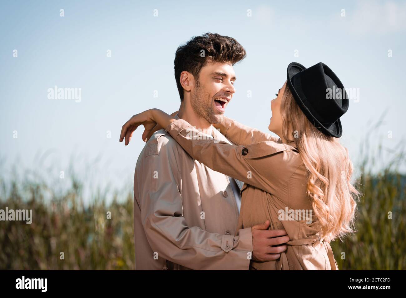 blonde woman in hat hugging excited man in trench coat Stock Photo - Alamy