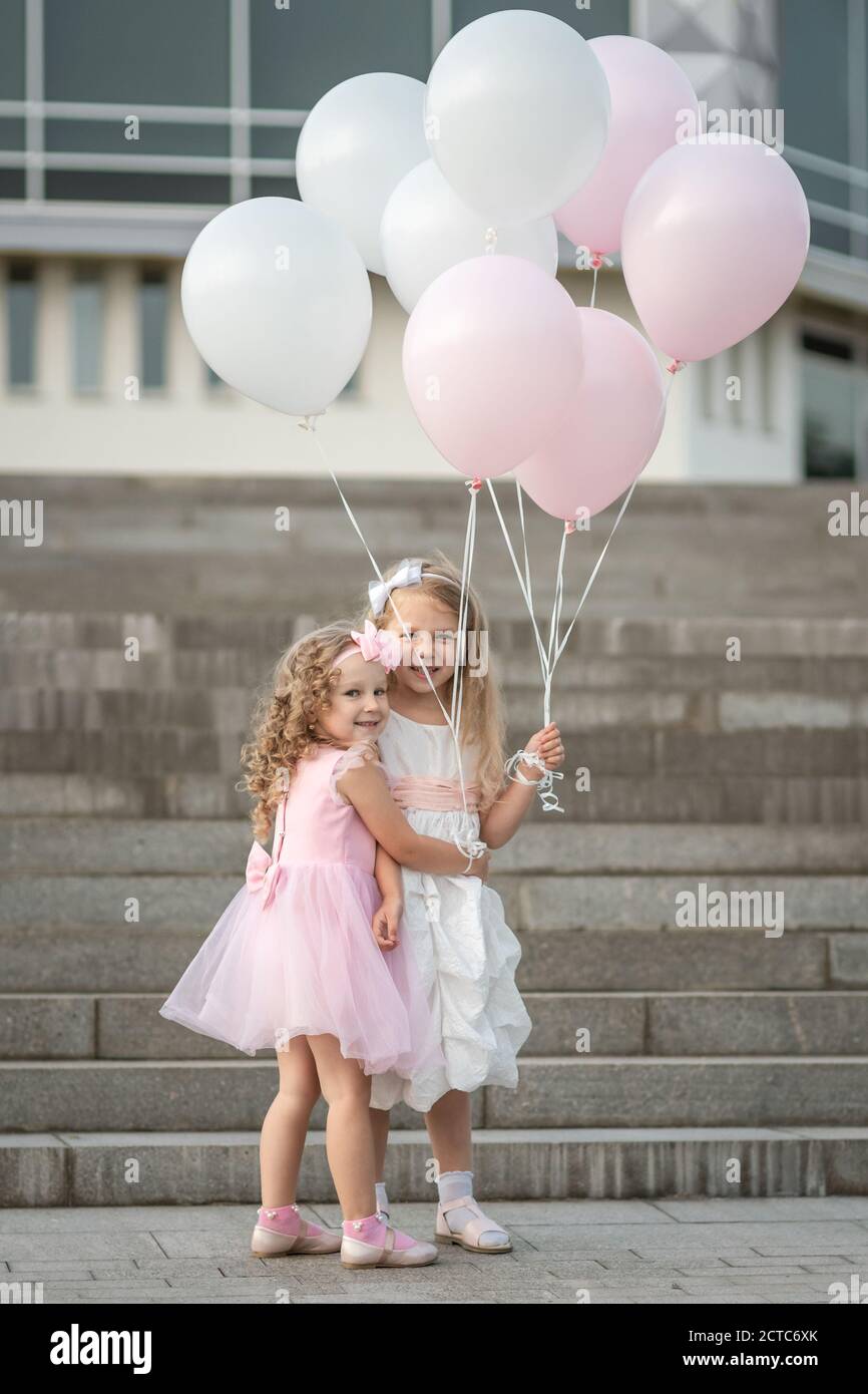Two little girls in pink and white dressses with balloons hugging on ...