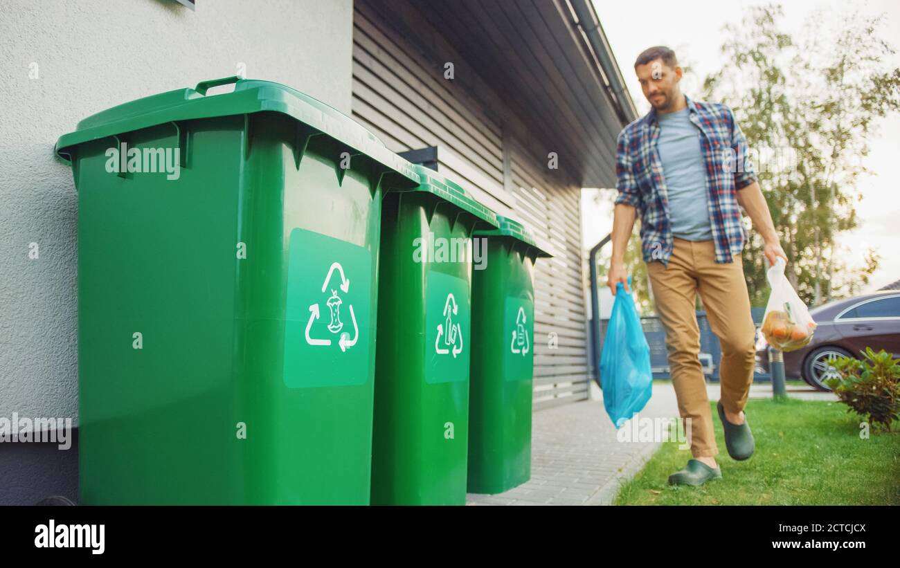 Caucasian Man is Walking Outside His House in Order to Take Out Two Plastic Bags of Trash. One Garbage Bag is Sorted with Biological Food Waste, Other Stock Photo