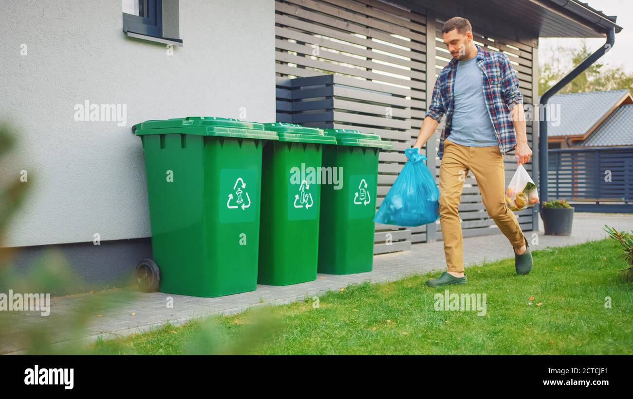 Caucasian Man is Walking Outside His House in Order to Take Out Two Plastic Bags of Trash. One Garbage Bag is Sorted as Biological Food Waste, Other Stock Photo