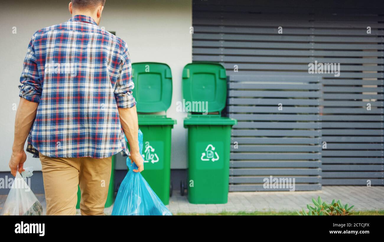 Caucasian Man is Walking Outside His House in Order to Take Out Two Plastic Bags of Trash. One Garbage Bag is Sorted with Biological Food Waste, Other Stock Photo