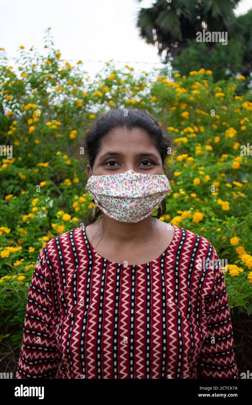 Beautiful lady with floral mask, earrings and designer top stands before yellow flowers. Protection from corona in a stylish manner. Stock Photo