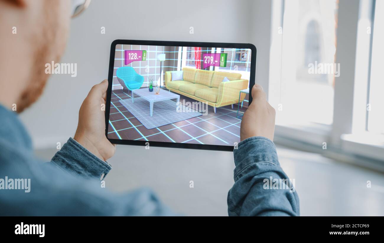 Decorating Apartment: Man Holding Digital Tablet with AR Interior Design Software Chooses 3D Furniture for Home from Online Shop with Shown Prices Stock Photo