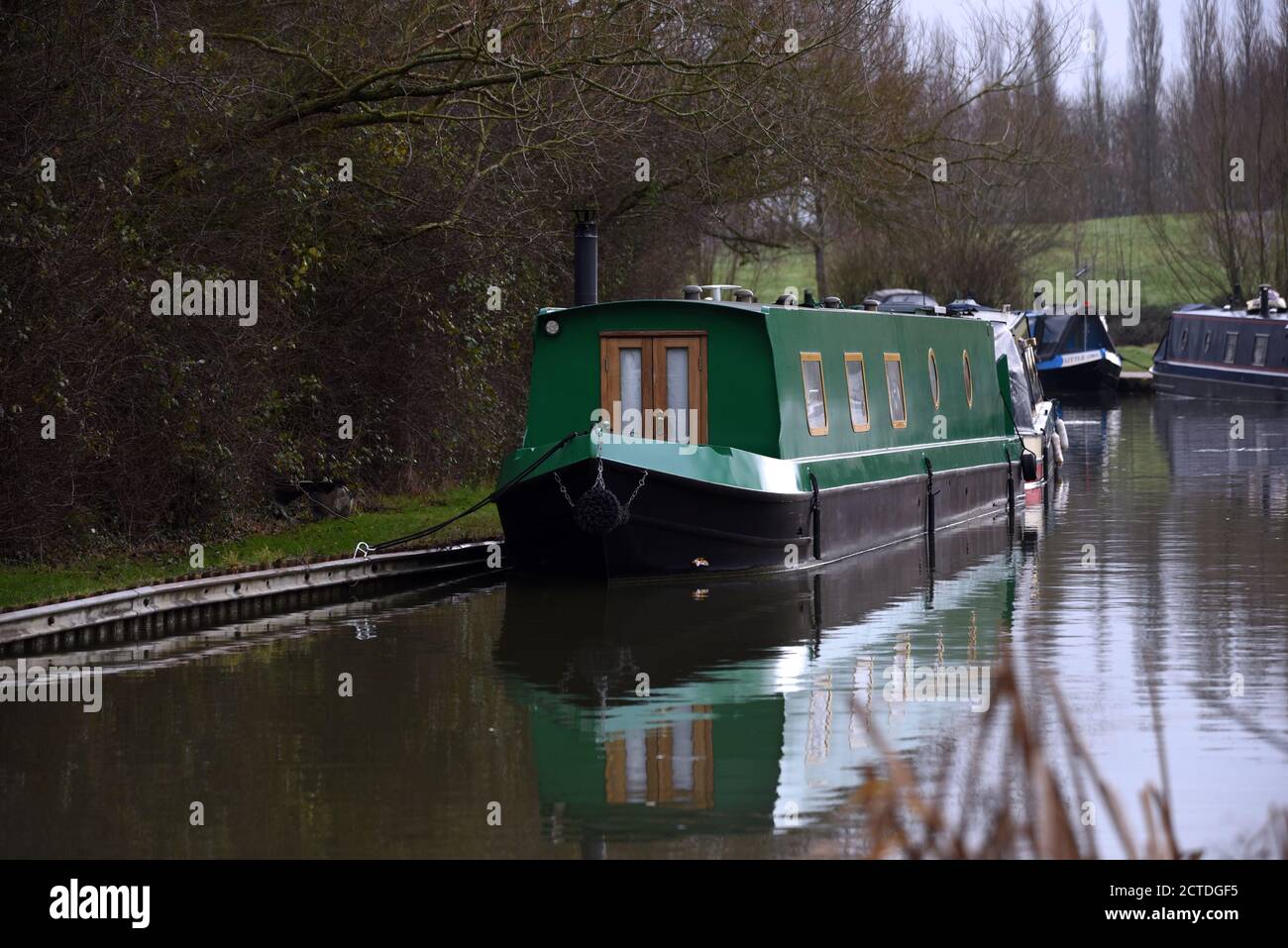A green canal boat is reflected in still water in this photo taken along the Grand Union canal in Buckinghamshire Stock Photo