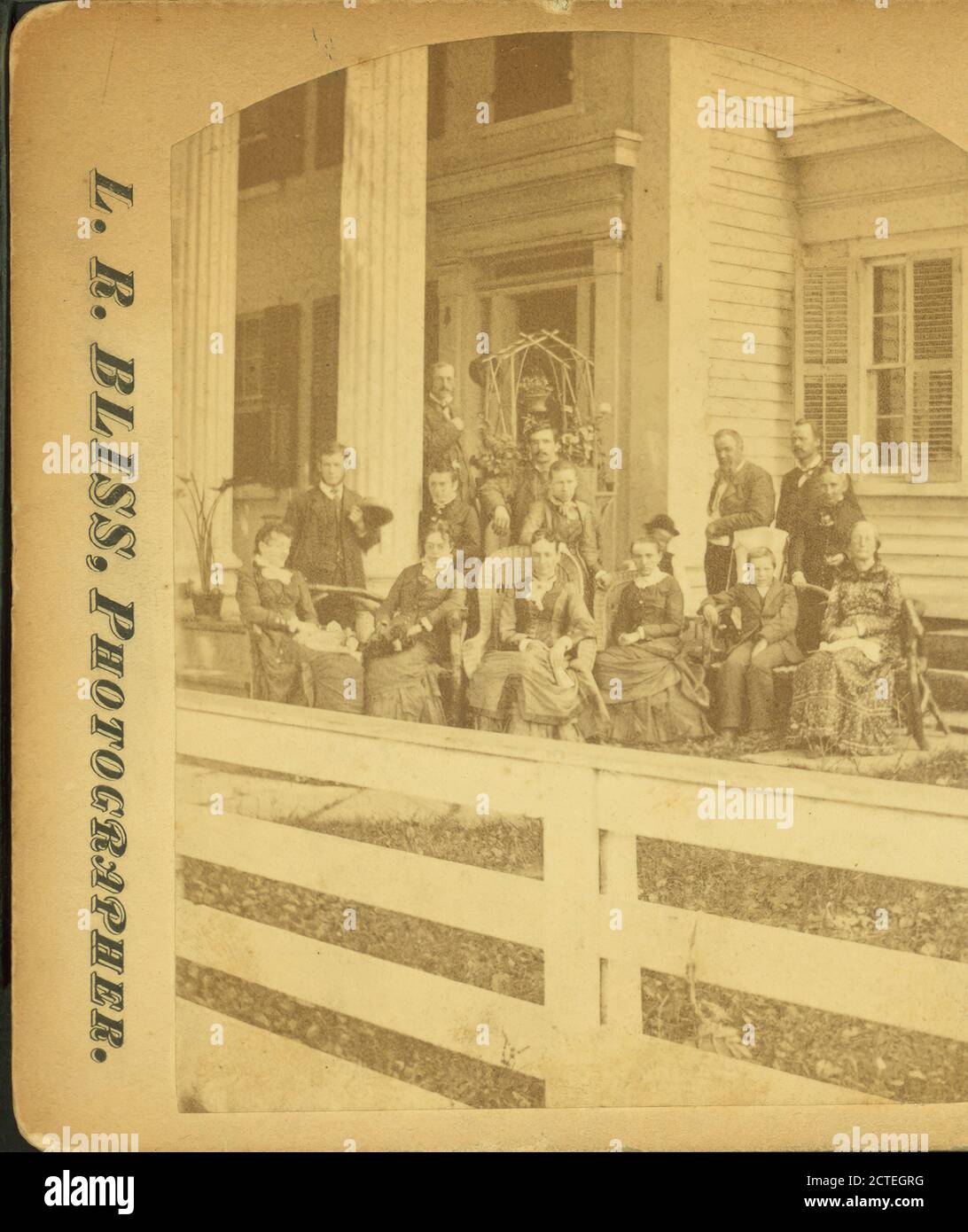 Multi-generation male and female family members, wearing suits and dark dresses, pose before white-columned porch and trellis of multi-story clapboard house; white board fence in foreground., Bliss, L. R., Pennsylvania Stock Photo