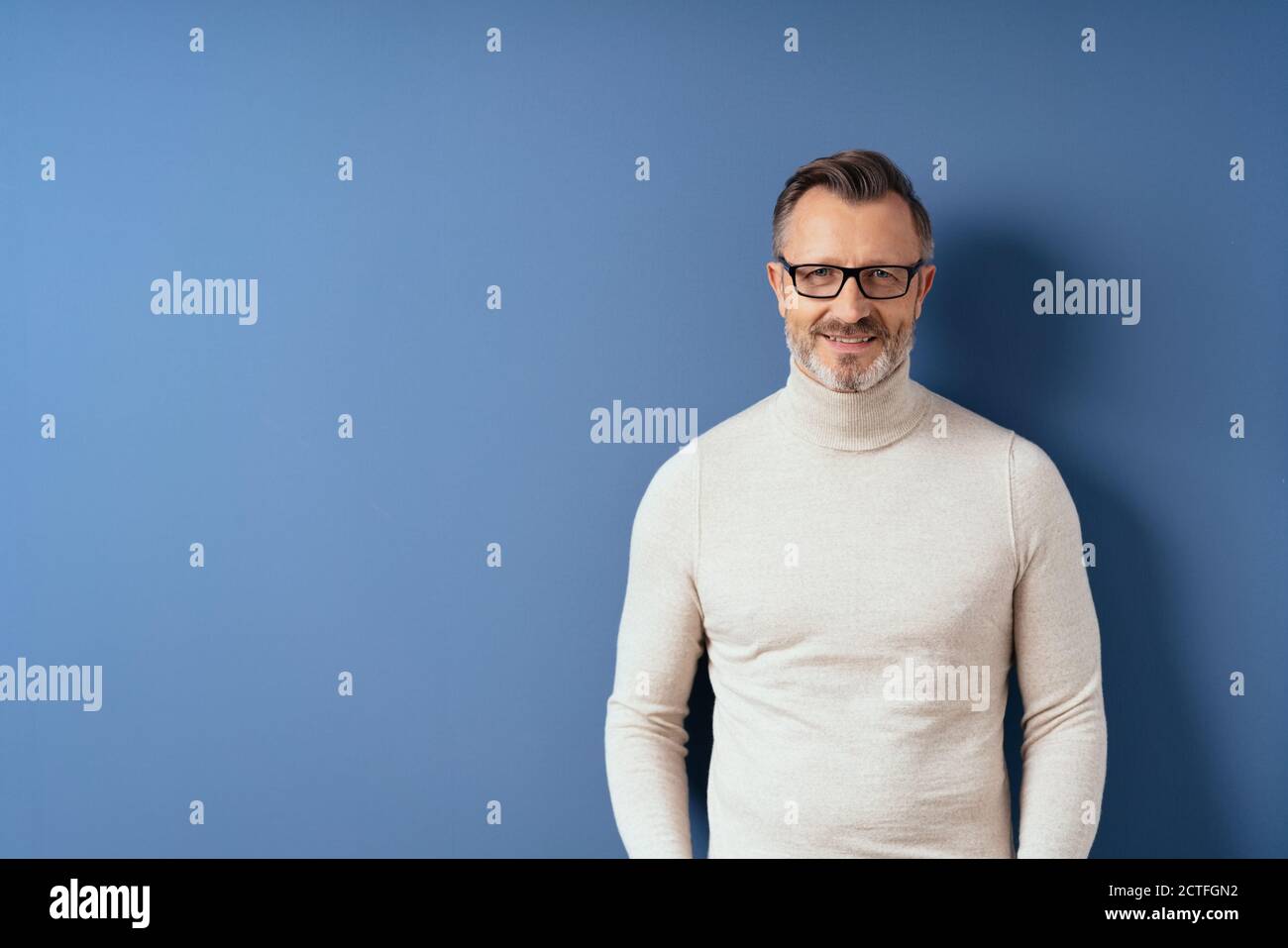 Half-length front portrait of a smiling handsome middle-aged man in white turtleneck against blue wall background with copy space Stock Photo