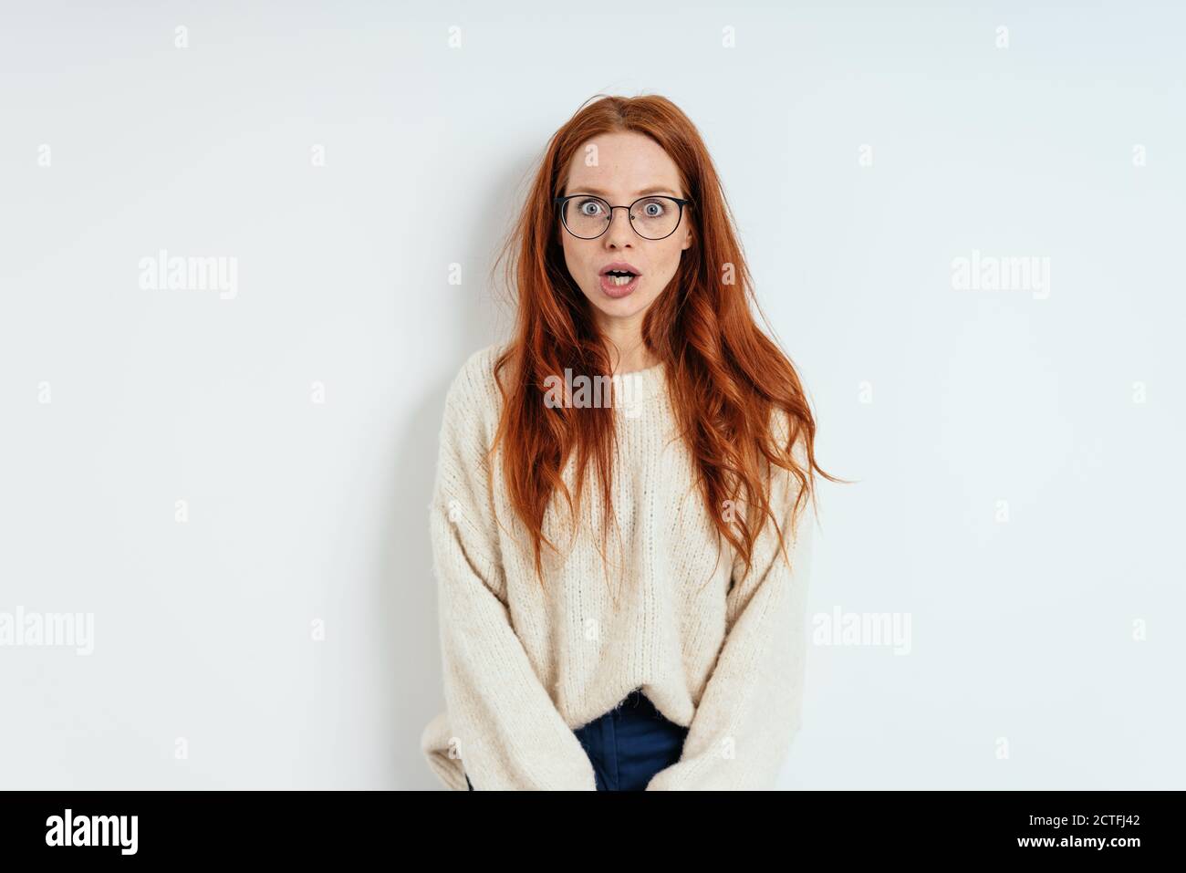 Shocked young woman in glasses with her mouth agape standing staring wide eyed at the camera over a white studio background with copy space Stock Photo
