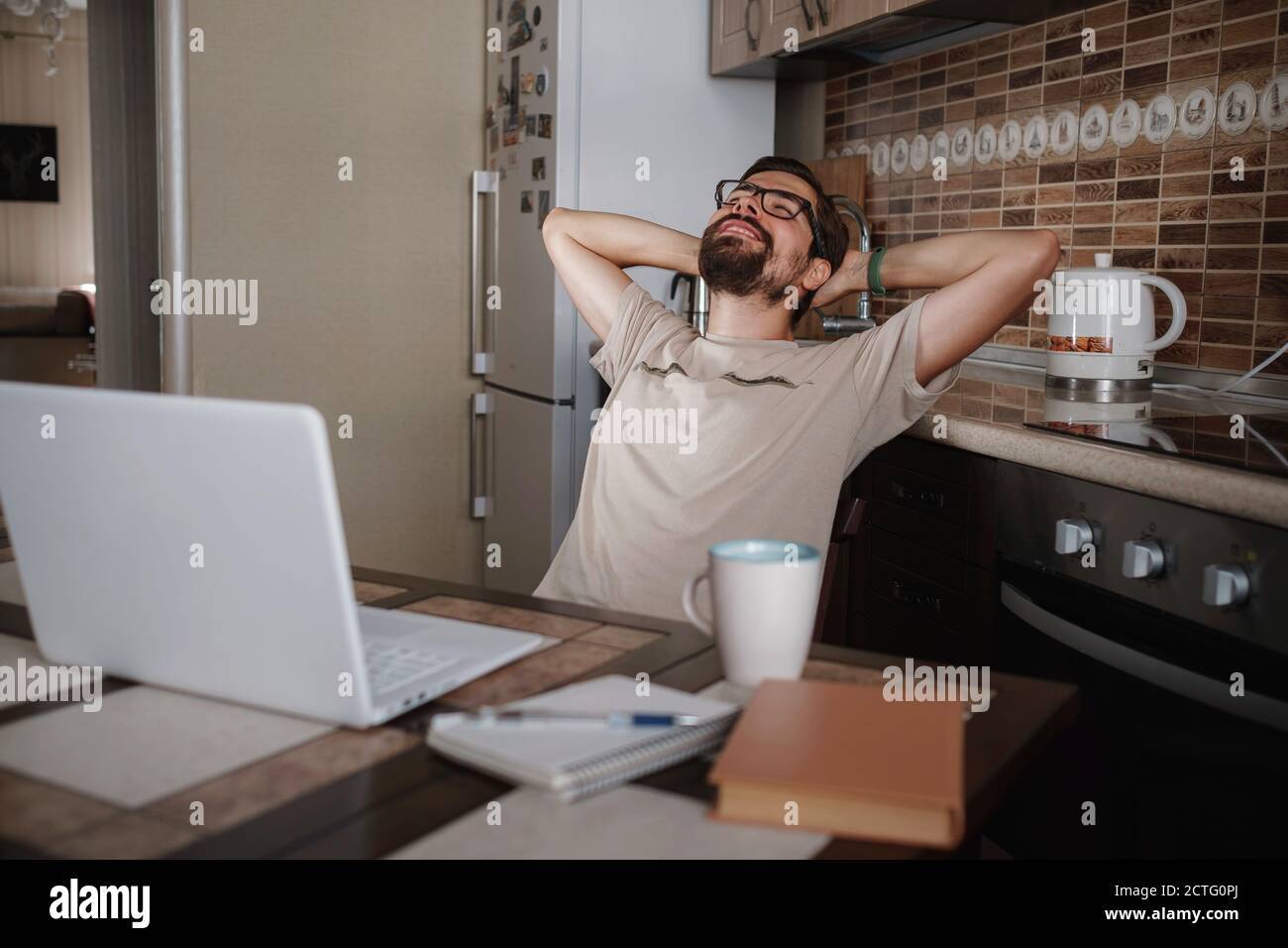 Smiling hipster freelancer holding hands behind head sitting at office desk behind laptop. Happy employee feeling no stress, relaxing, watching funny Stock Photo