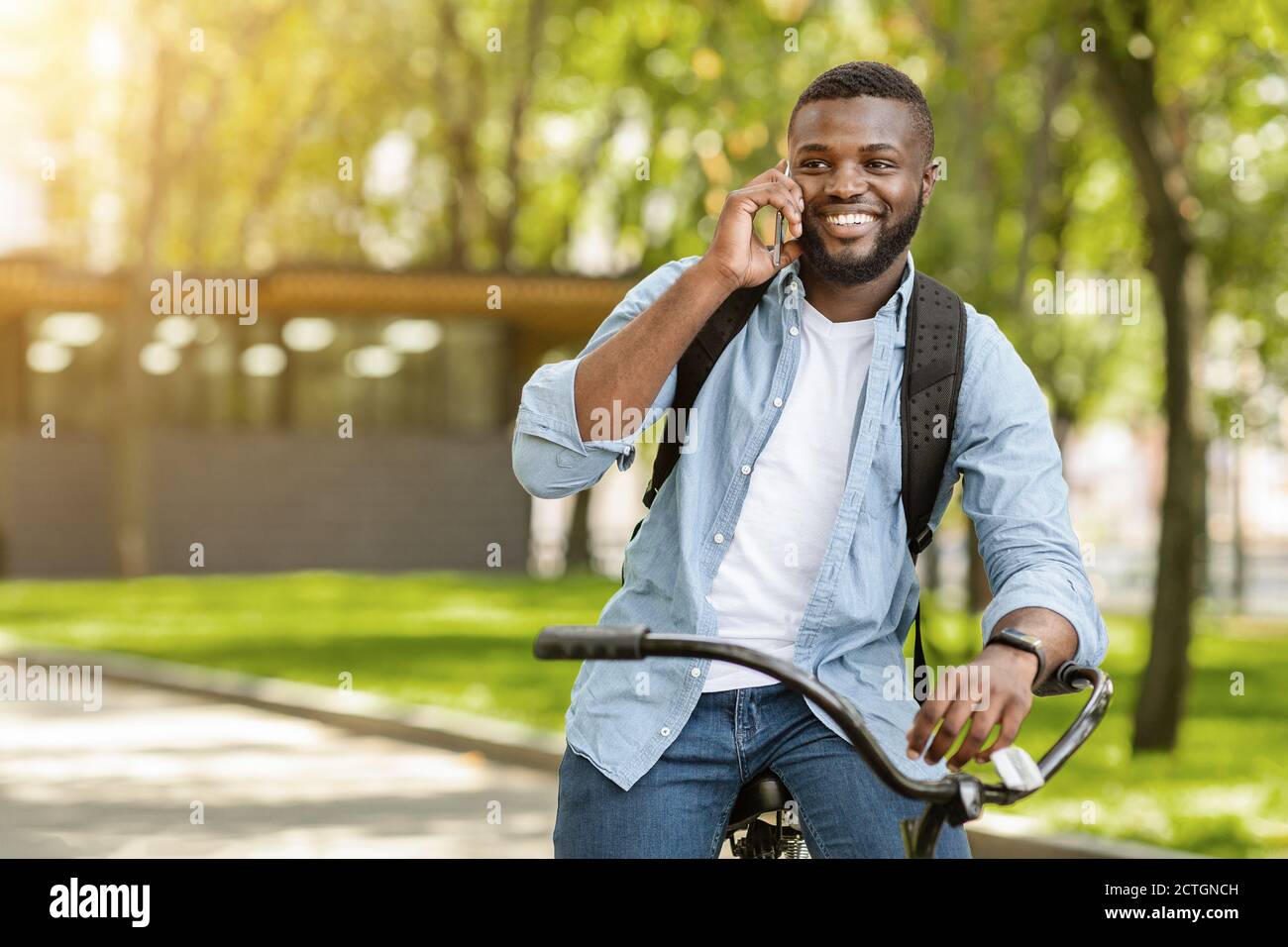 Smiling Black Guy Talking On Cellphone While Riding Bicycle Outdoors Stock Photo