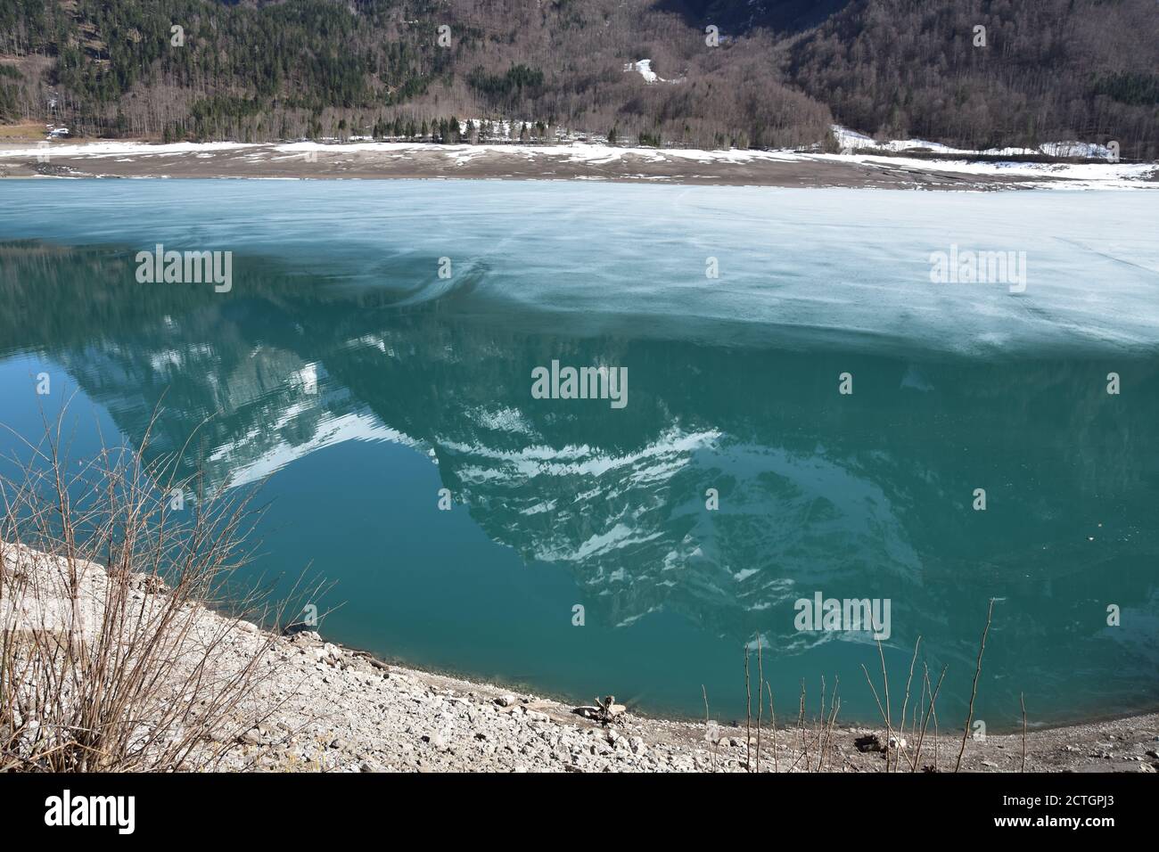 Half-empty Klontalersee lake with reflection of Alps in the water and still covered partly with ice in Klontal, Kloental valley, during sunny day. Stock Photo
