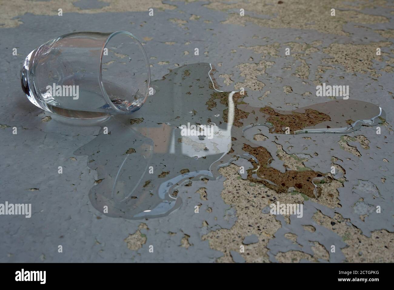 Glass of water overturned or tipped over on the concrete plate with peeling off old paint. The spilled liquid forms irregular puddle around the glass. Stock Photo