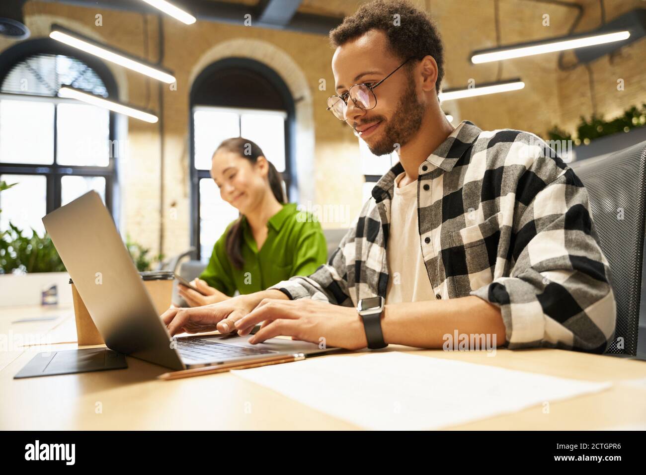Young smart guy wearing eyeglasses working on laptop while sitting at desk with his female colleague in the modern coworking space or office. Business people and teamwork concept Stock Photo
