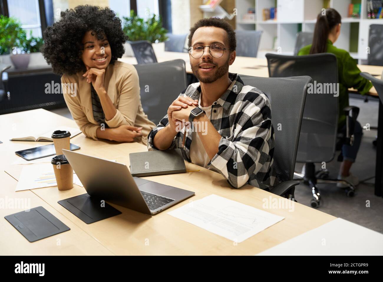 Young happy mixed race guy wearing eyeglasses looking at camera and smiling, sitting at desk with his cheerful female colleague in the modern coworking space. Business people working together Stock Photo