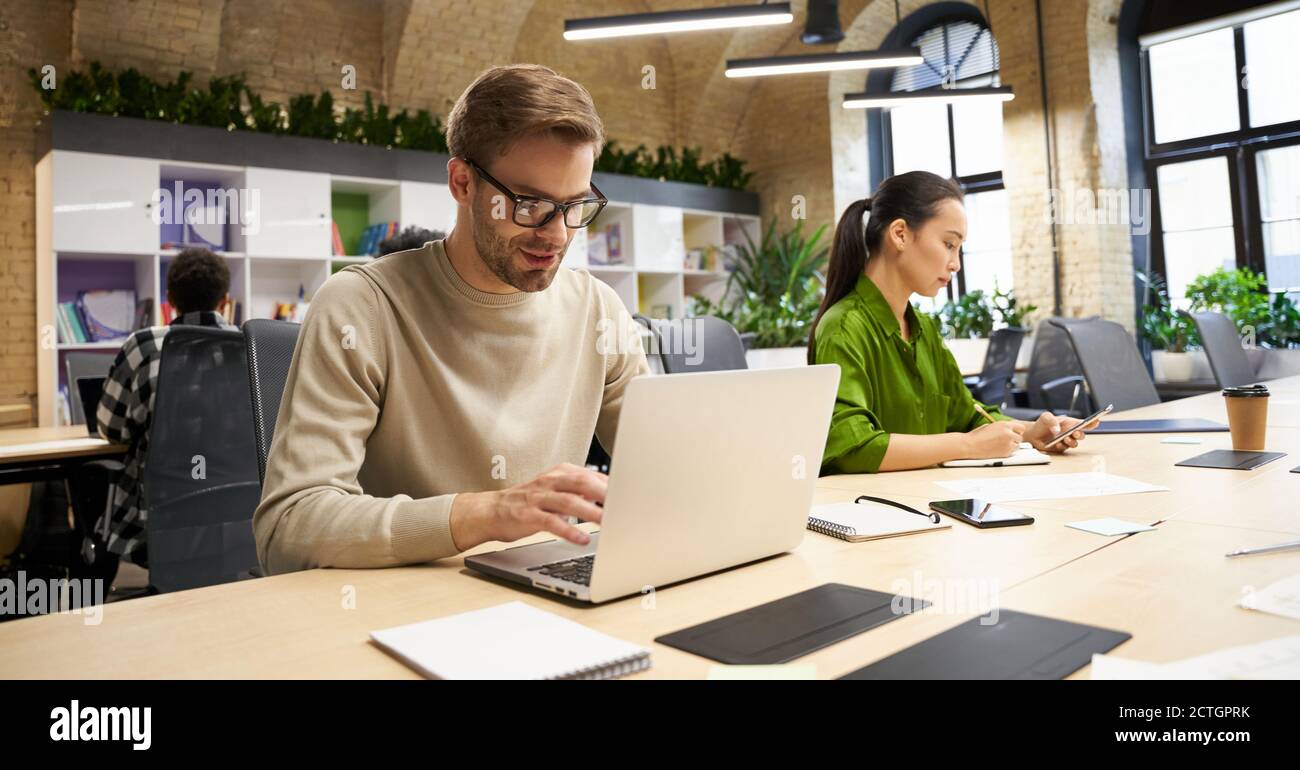 Young focused caucasian man wearing eyeglasses working on laptop while sitting at desk with his female colleague in the modern coworking space. Business people and teamwork concept Stock Photo