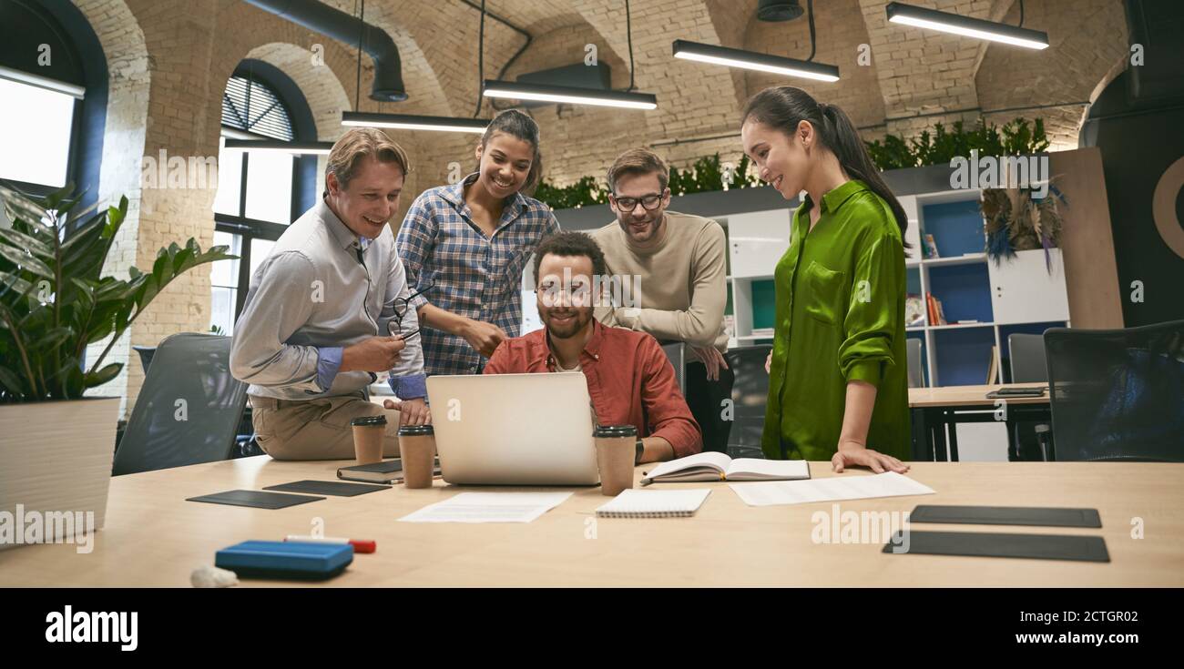 Business professionals. Multicultural team, group of young cheerful business people analyzing data, looking at laptop screen while working together in the office. Project team in coworking space Stock Photo