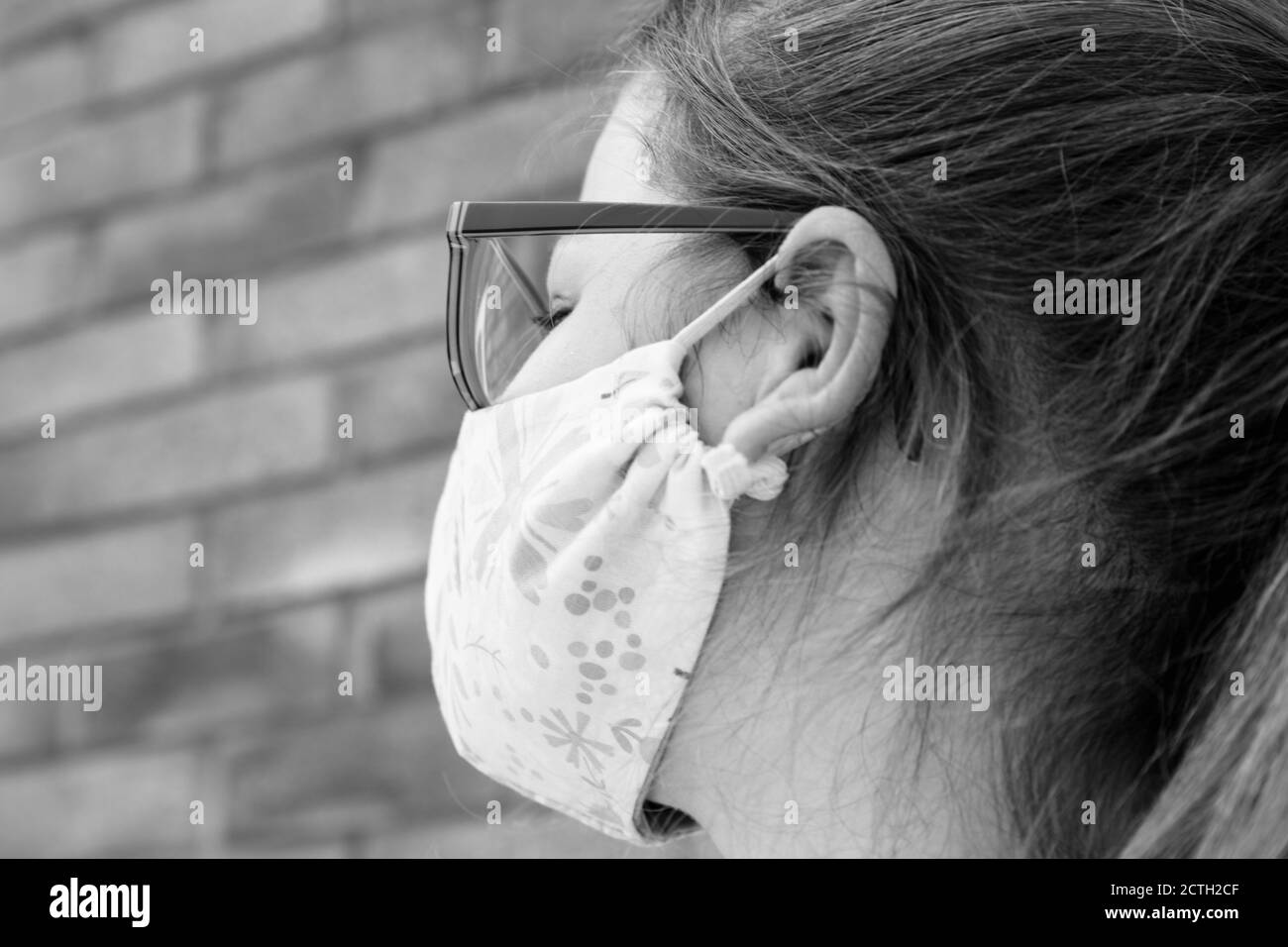 close up of little girl's face wearing pretty fabric face mask outside, against a brick wall background in summer sun Stock Photo