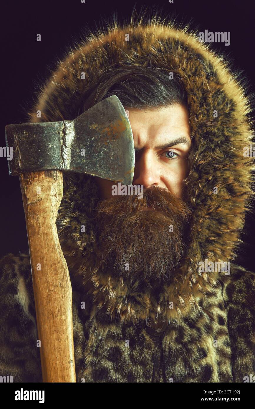 handsome bearded man or woodman guy with fashionable mustache and beard on serious face in brown fur coat, holds sharp axe or ax on black background Stock Photo