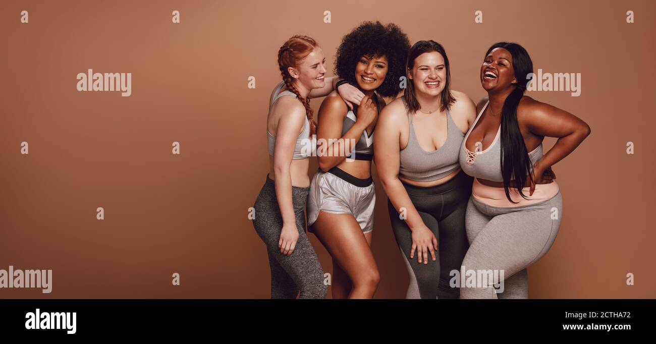 Smiling group of women in different size standing together in sportswear against brown background. Diverse group women looking at camera and laughing. Stock Photo
