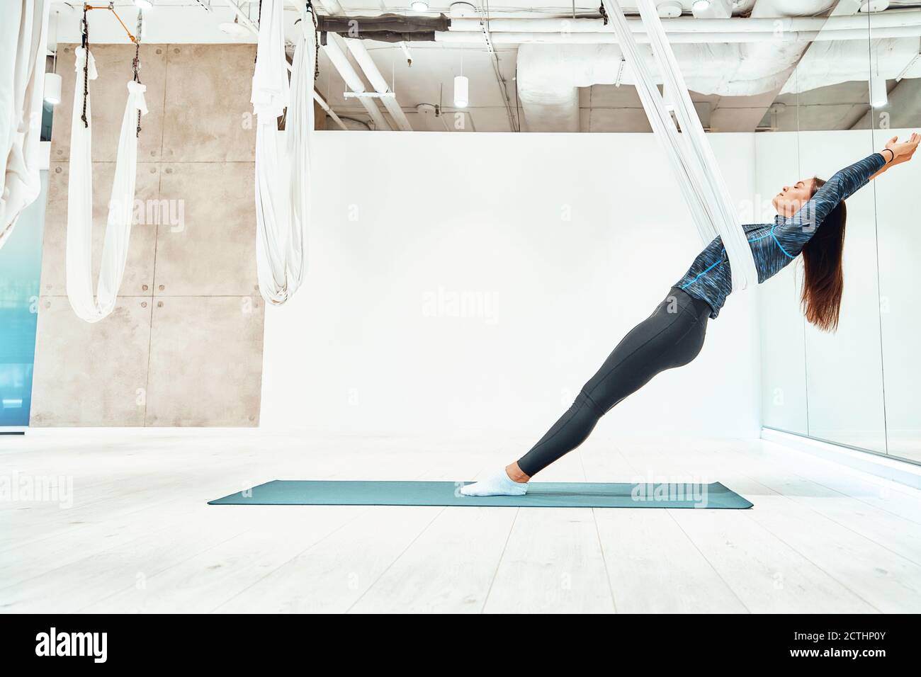 Young caucasian woman in sportswear practicing fly yoga in beautiful bright studio. Sport, wellness and healthy lifestyle, aerial yoga concept Stock Photo
