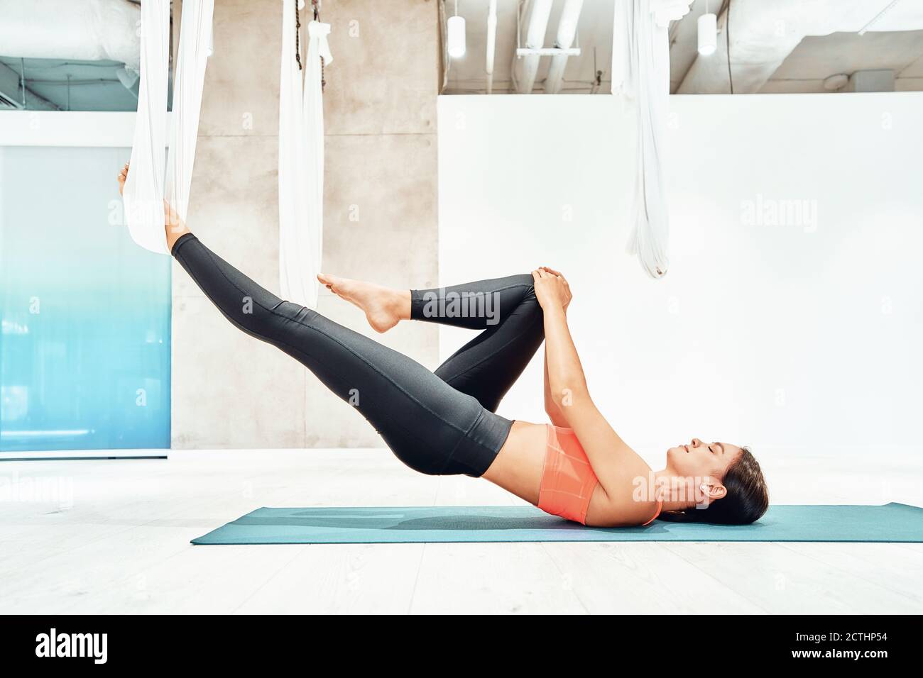 Young caucasian fitness woman in sportswear practicing fly yoga in studio, stretching body with white hammock or swing suspended from the ceiling. Sport, wellness and healthy lifestyle, aerial yoga Stock Photo