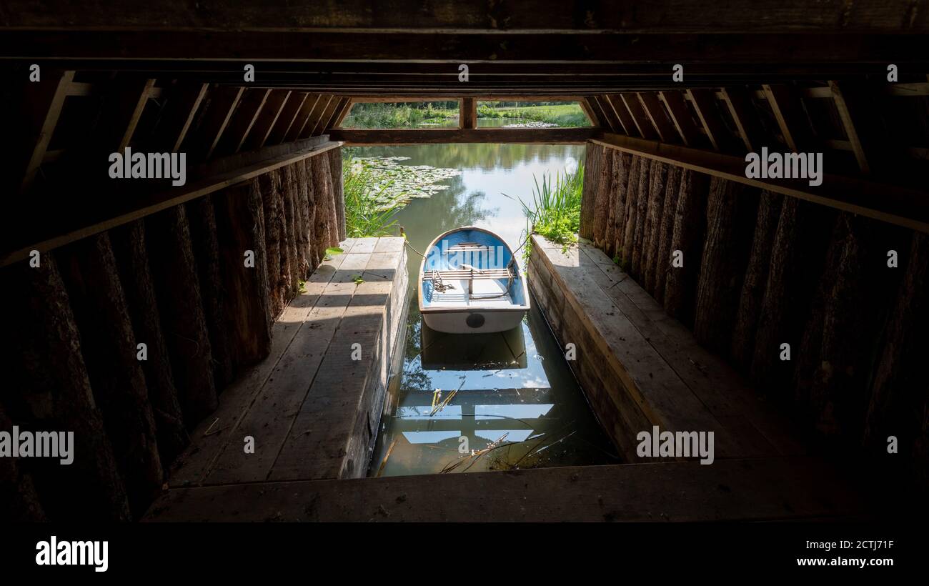 A small white and blue rowing boat docked up inside a small wooden shack at the side of a lake at Arundel Castle Stock Photo