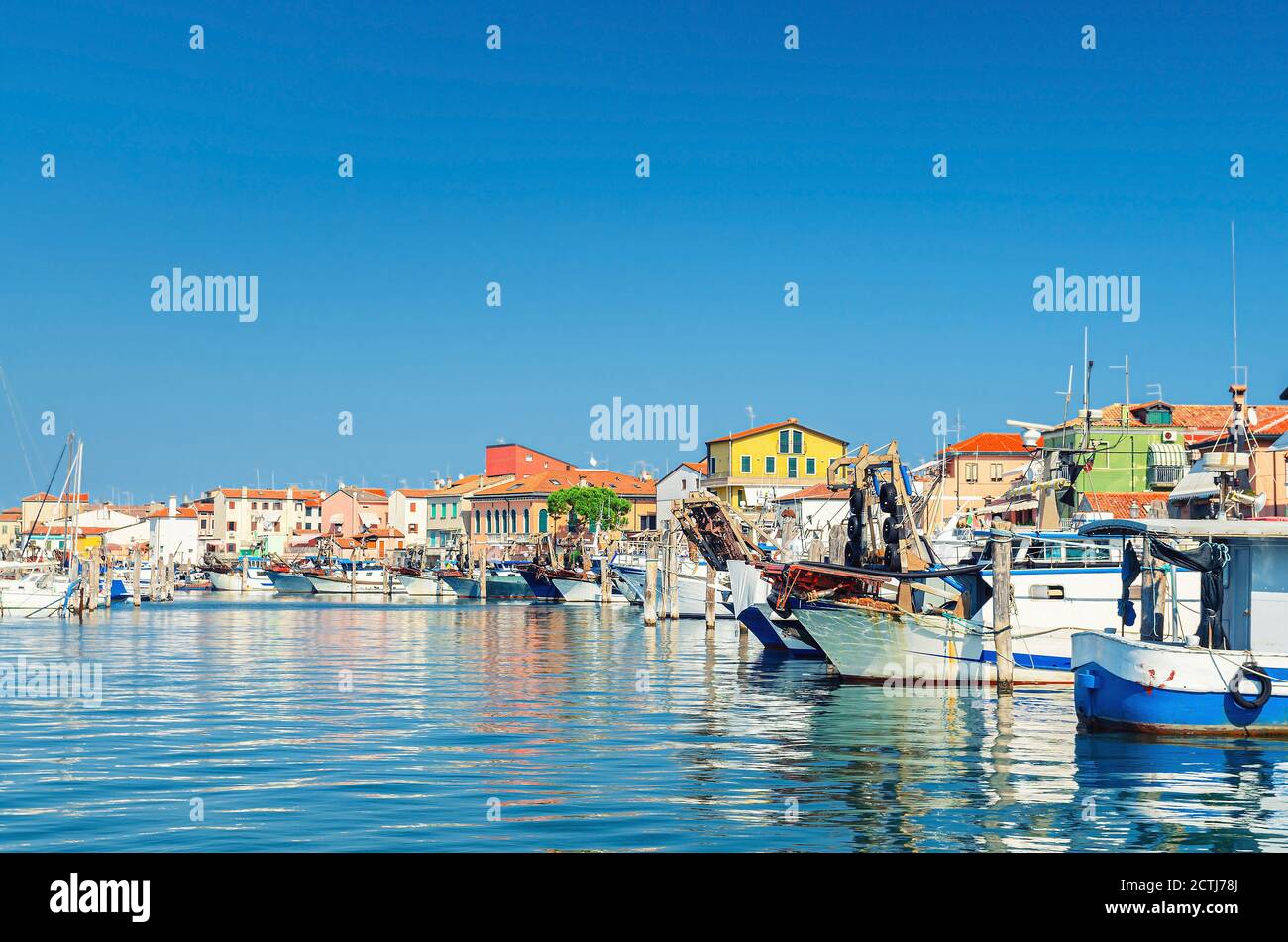 Fishing ships and boats moored in port lagoon near seaside of Sottomarina town with row of colorful buildings in summer day, blue sky background, Veneto Region, Northern Italy Stock Photo