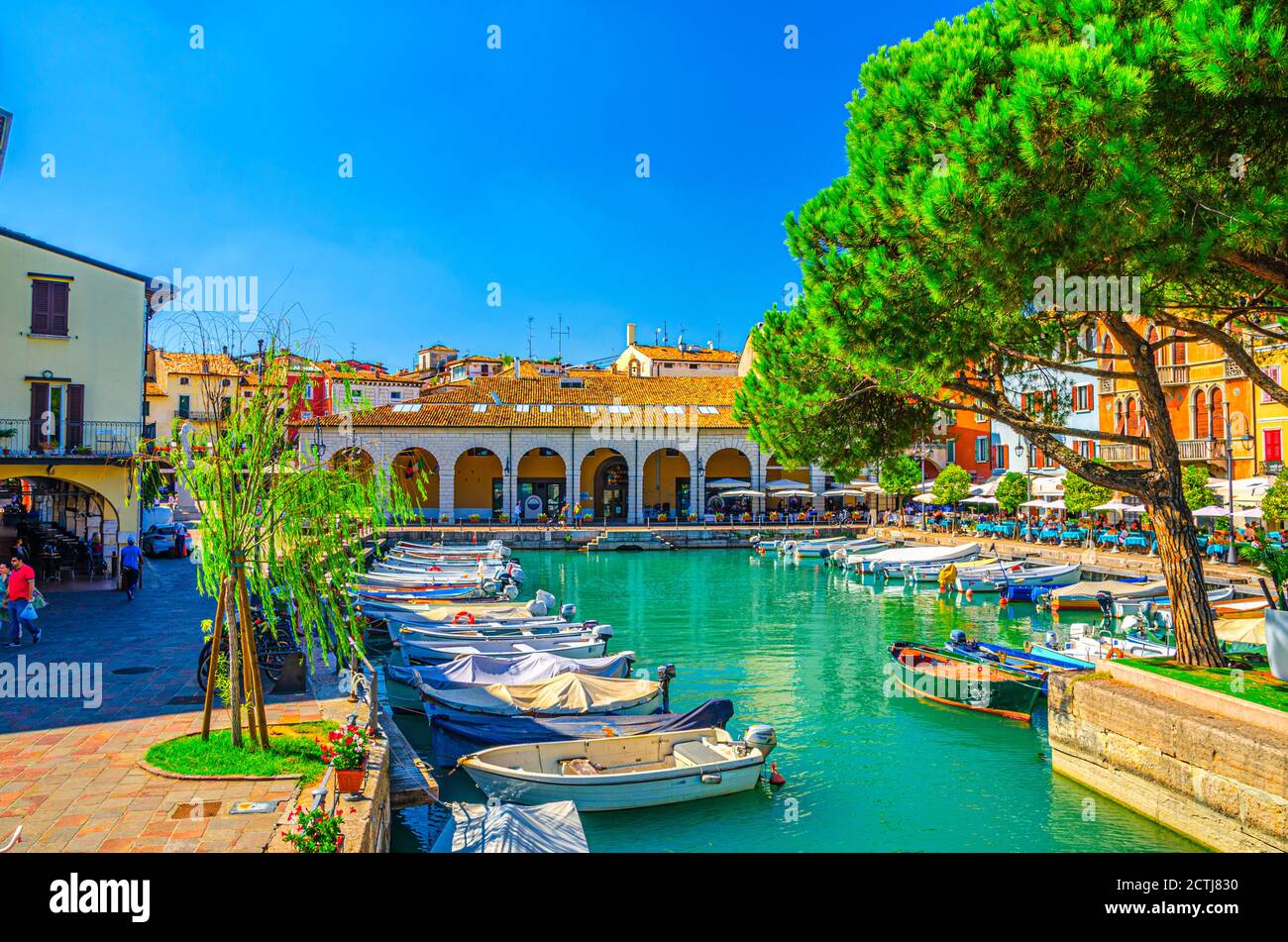 Desenzano del Garda, Italy, September 11, 2019: Old harbour with boats on turquoise water, green trees, street restaurants and traditional buildings in historical town centre, blue sky, Lombardy Stock Photo