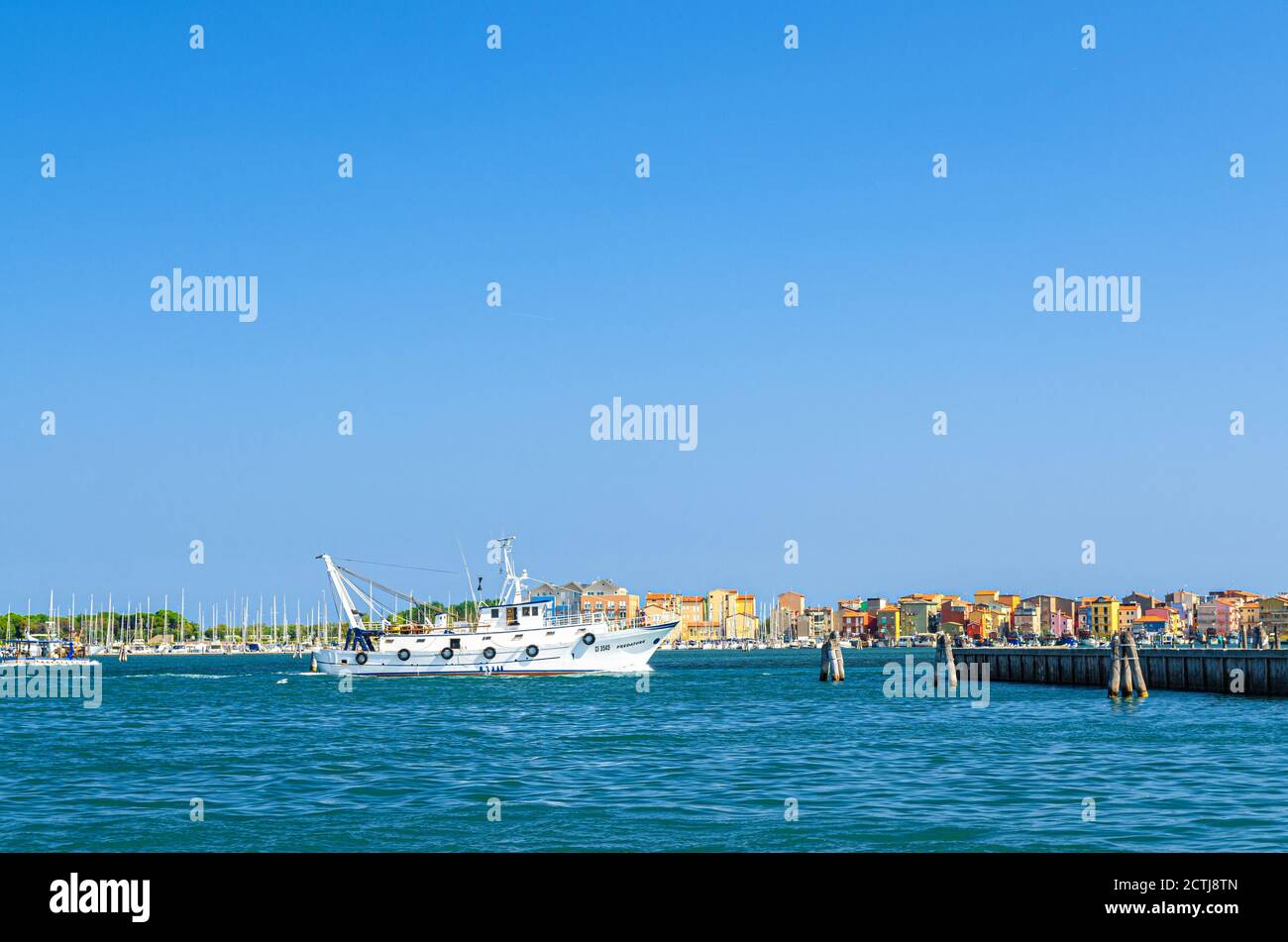Chioggia, Italy, September 16, 2019: fishing ship boat in water of lagoon with row of colorful buildings of Sottomarina town, blue sky background in summer day, Veneto Region Stock Photo
