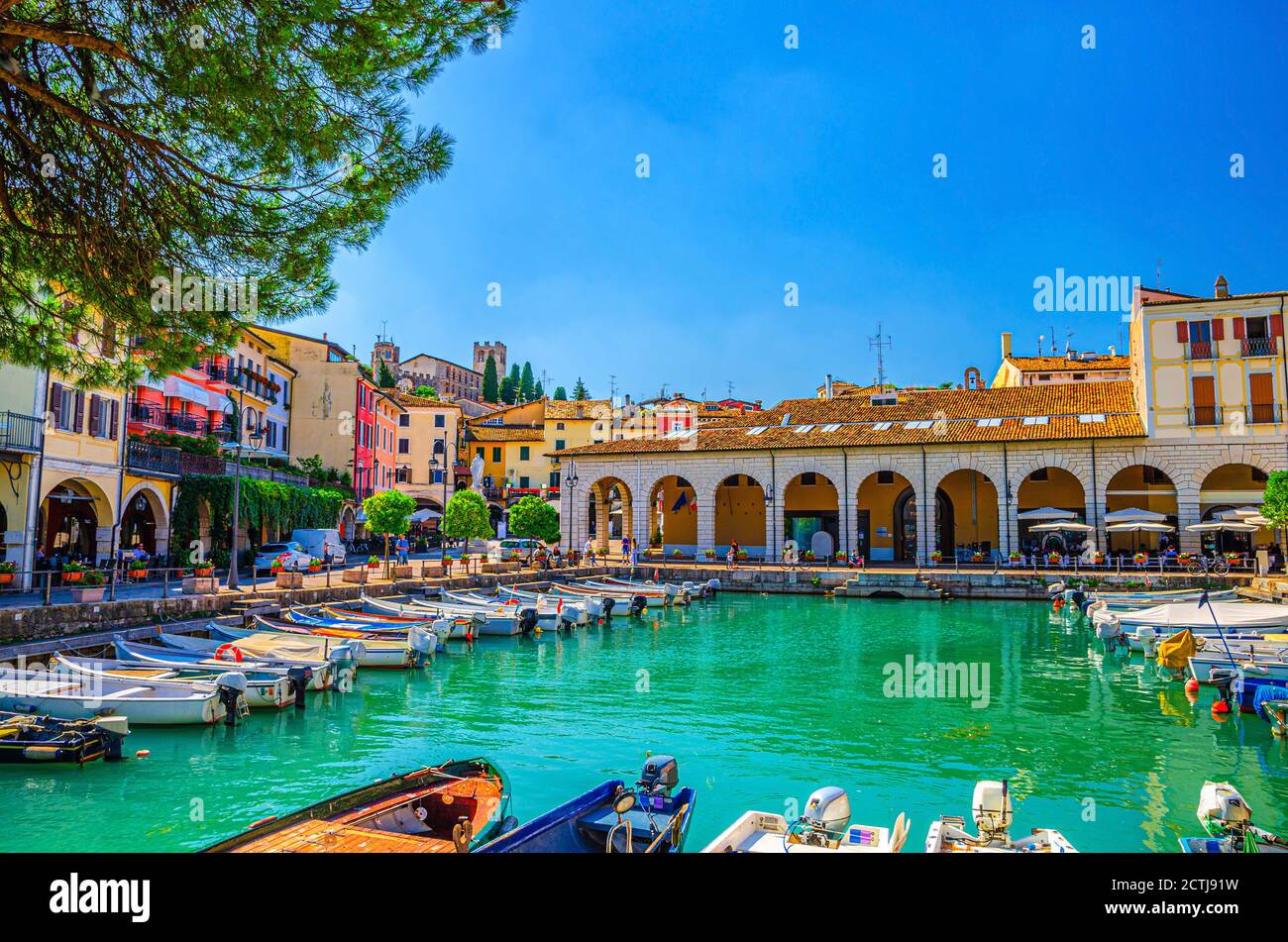 Old harbour Porto Vecchio with motor boats on turquoise water, green trees and traditional buildings in historical centre of Desenzano del Garda town, blue sky background, Lombardy, Northern Italy Stock Photo
