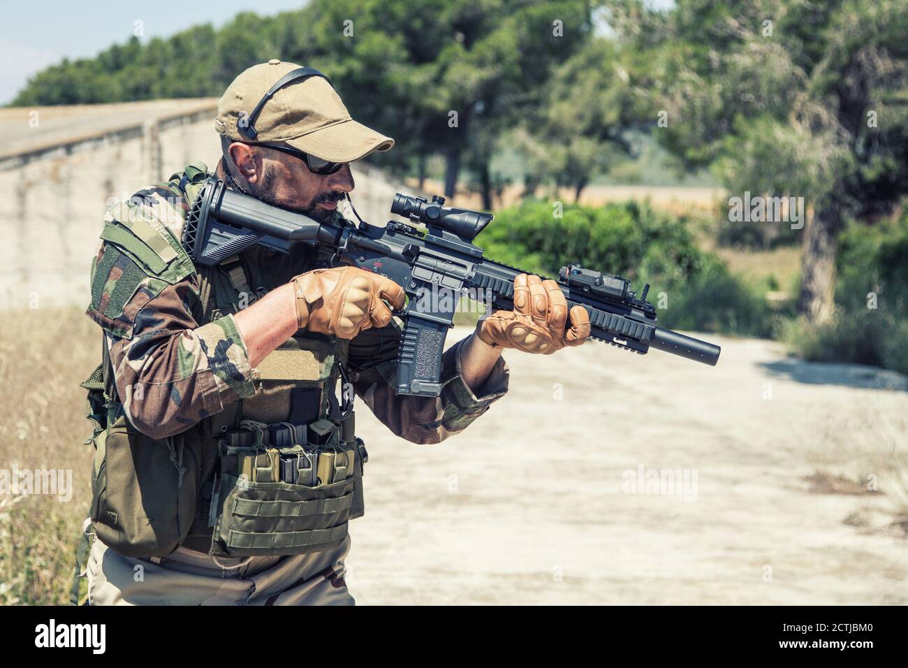 Private military company mercenary, brutal looking special forces fighter in battle uniform and plate carrier, wearing radio headset and sunglasses, holding service rifle in hands, ready to fight Stock Photo