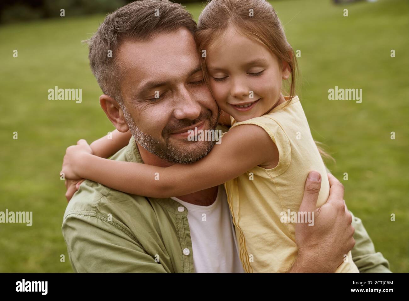 Portrait of young loving father and his cute little girl keeping eyes closed and hugging while spending time outddors, sitting on a green grass Stock Photo