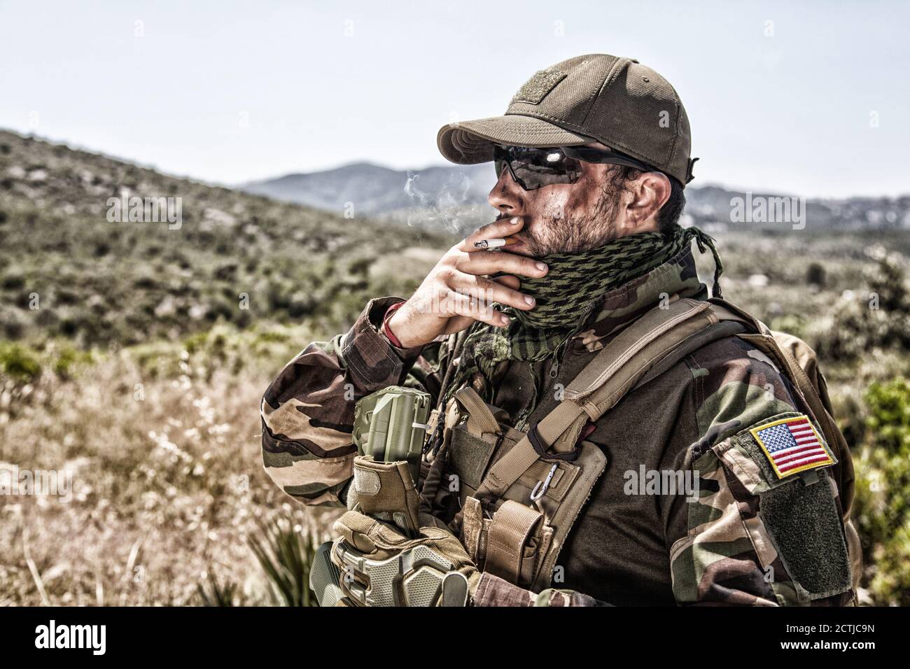 Army soldier, commando shooter in ammunition and battle uniform, standing in mountain area, looking into distance and smoking cigarette. Special operations forces fighter during mission Stock Photo