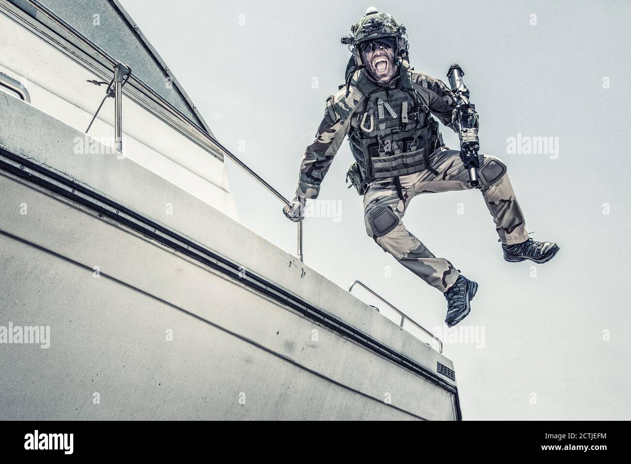 Army commando, special forces soldier equipped with body armour and battle helmet with radio headset, screaming and yelling while jumping from speed boat deck, landing on shore with assault rifle Stock Photo