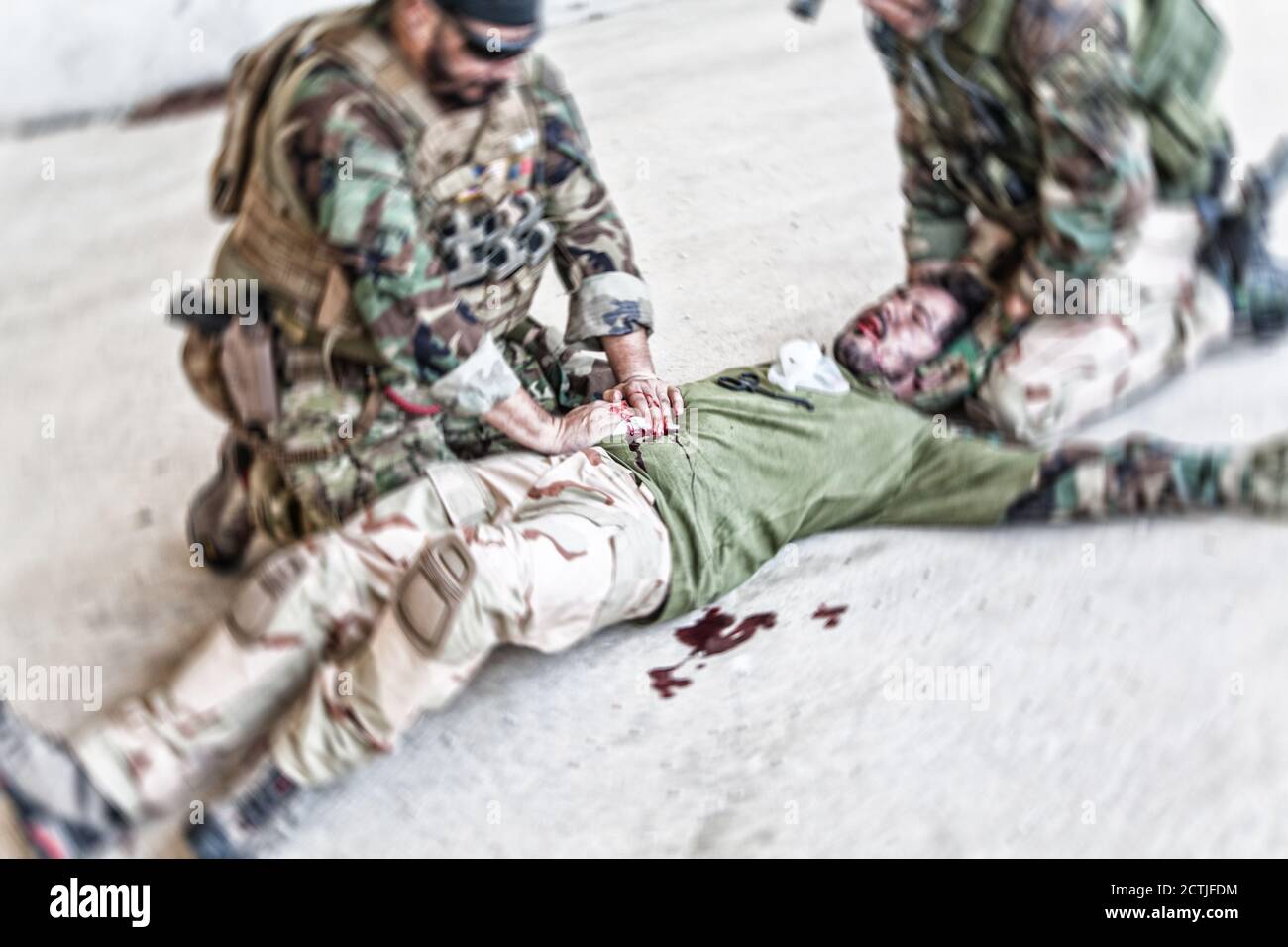 Soldiers trying to stop bleeding at wounded comrade who lying on floor, suffering and screaming in paine. Commando fighter pressing with hands on bloody wound at friends stomach, giving emergency care Stock Photo