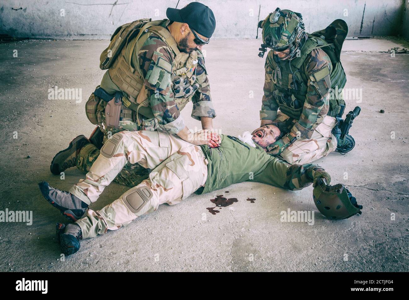 Soldiers trying to stop bleeding at wounded comrade who lying on floor, suffering and screaming in paine. Commando fighter pressing with hands on bloody wound at friends stomach, giving emergency care Stock Photo