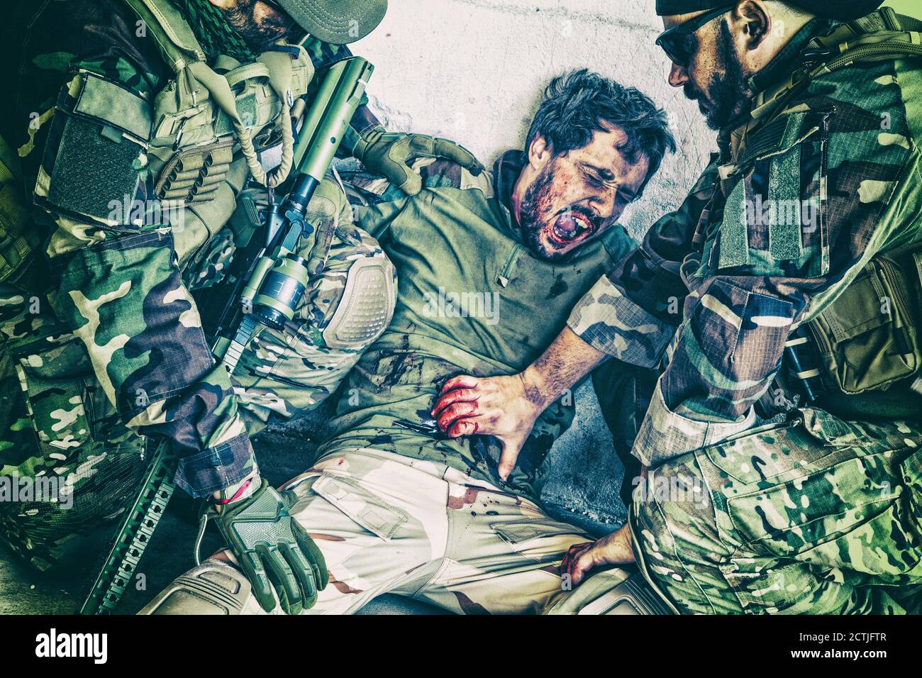 Commando fighters, Navy SEALs infantrymen giving emergency help to wounded comrade on battlefield. Soldier screaming in pain while teammate trying to stop bleeding from gunshot wound in his stomach Stock Photo