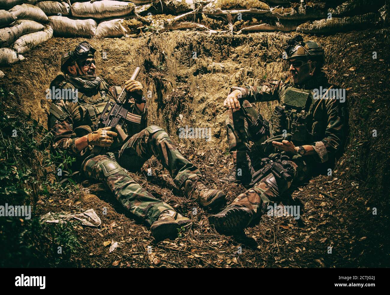 Smiling US army soldier, SEALs fighter, modern combatant in combat uniform, plate carrier, ballistic glasses and battle helmet resting after fight, sitting in trench at night and smoking cigarette Stock Photo