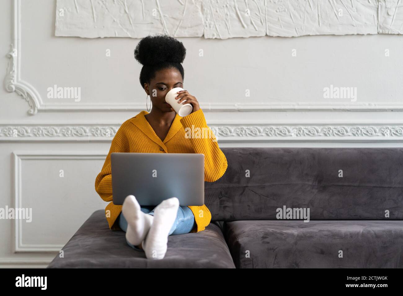 Afro-American biracial woman with afro hairstyle in yellow cardigan sitting on couch, working online on laptop, watching webinar or videos in social m Stock Photo