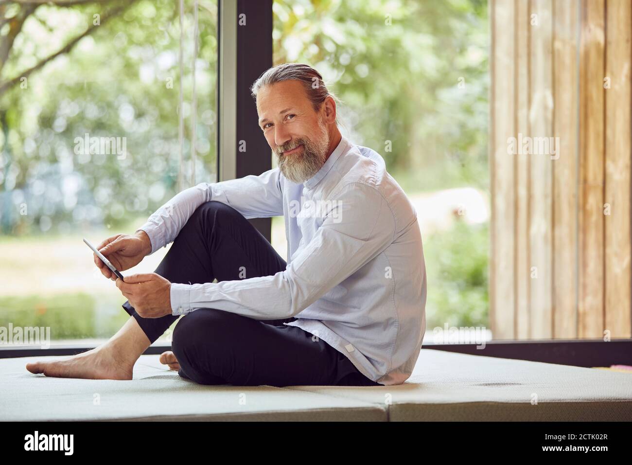 Smiling bearded man using digital tablet while relaxing on bed by window at home Stock Photo