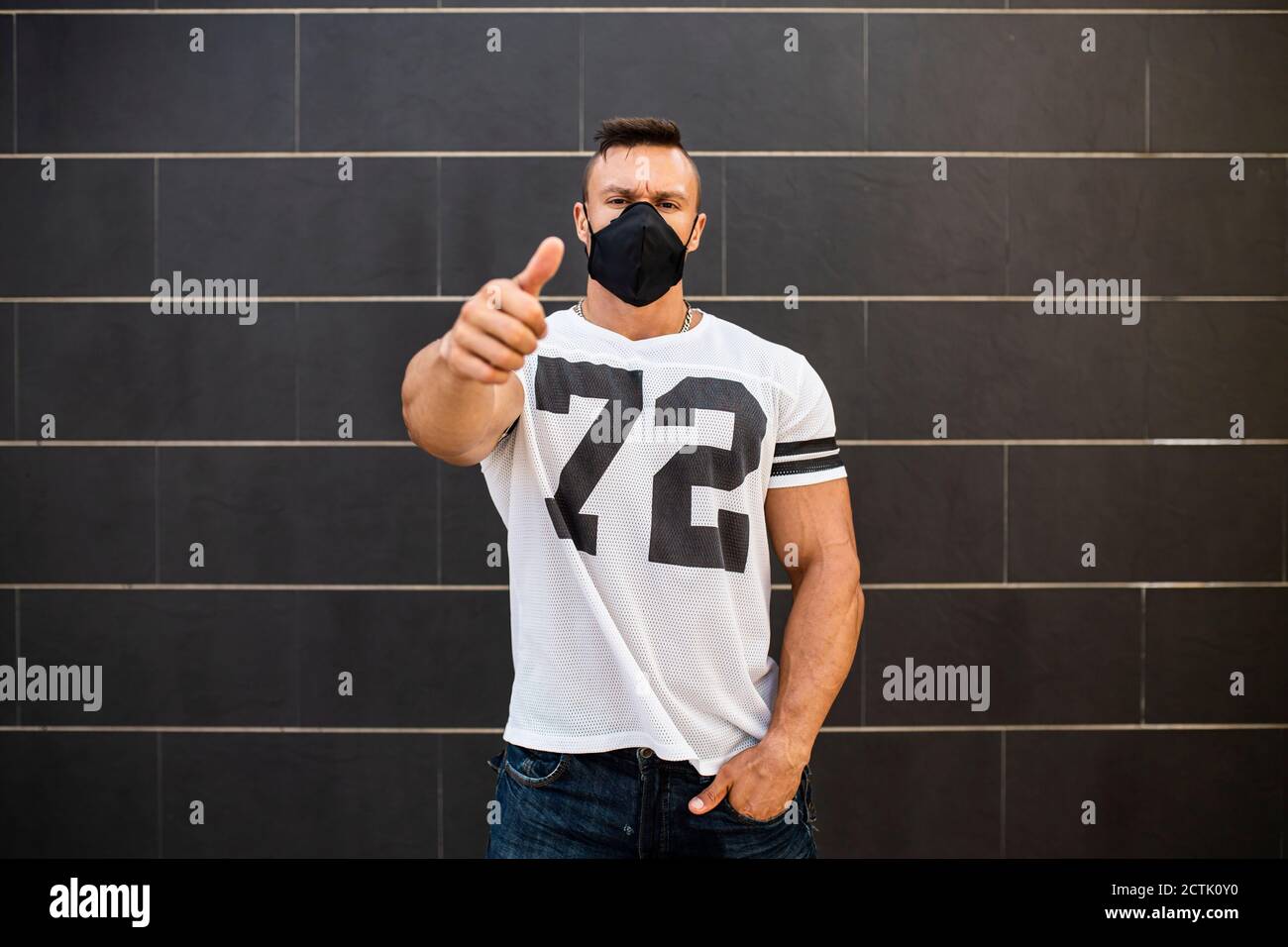 Macho man wearing face mask showing thumbs up while standing against wall Stock Photo