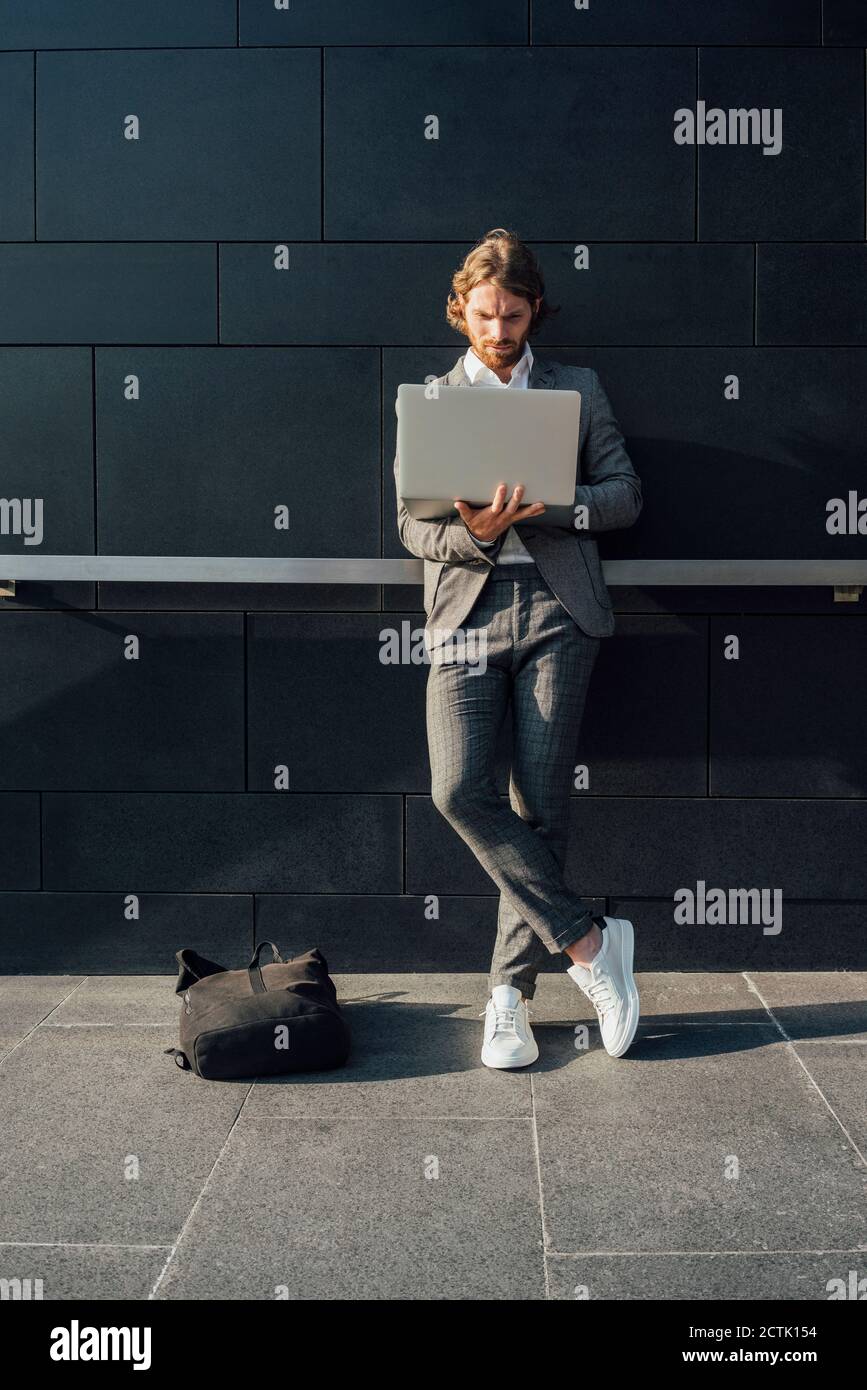Male entrepreneur using laptop while standing with crossed legs against wall on sunny day Stock Photo