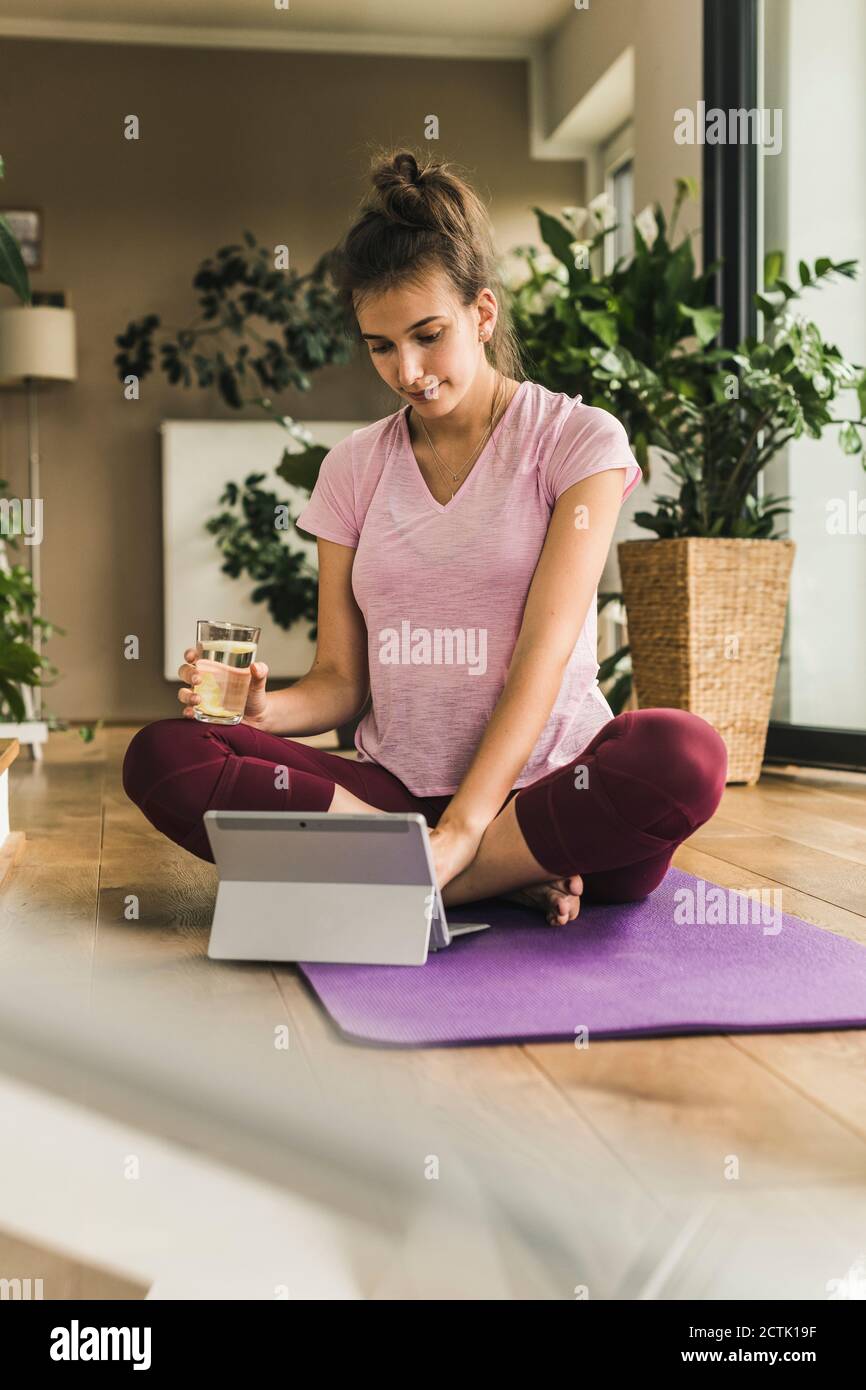 Young woman holding water using digital tablet while sitting on exercise mat at home Stock Photo