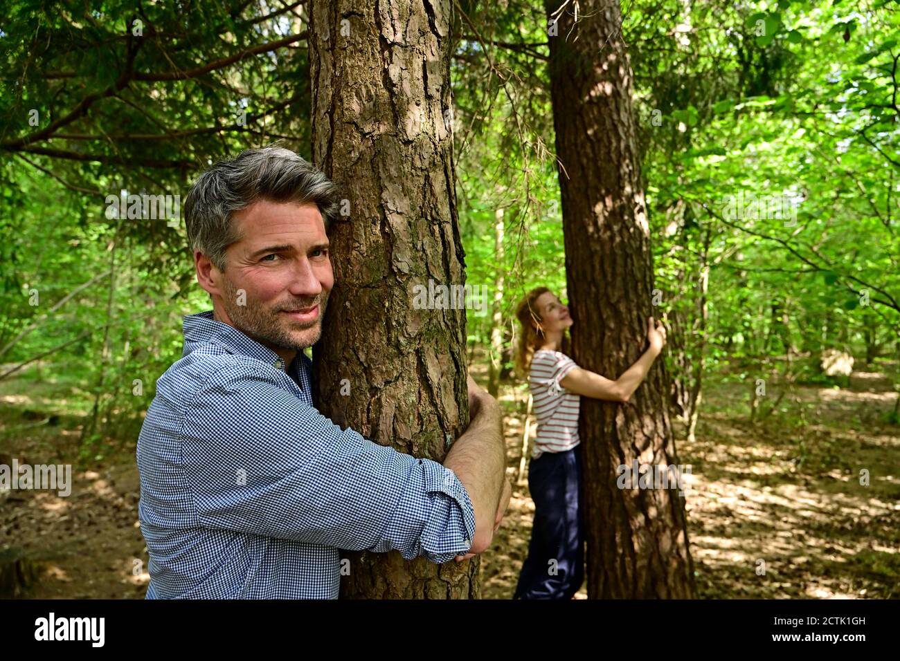 Smiling man and woman hugging tree while standing in forest Stock Photo ...