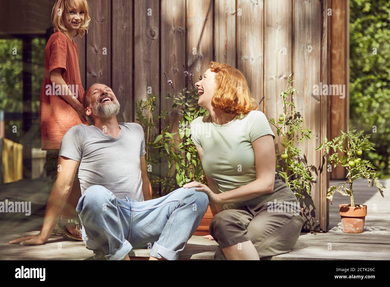 Girl standing by cheerful parents laughing while sitting outside tiny house Stock Photo