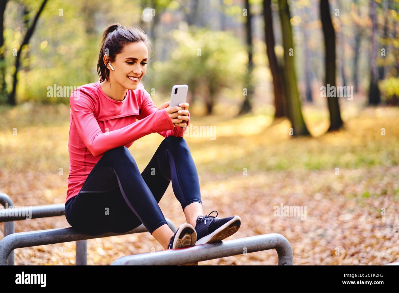 Young female jogger using smartphoneon sitting on bicycle stand in autumn forest Stock Photo