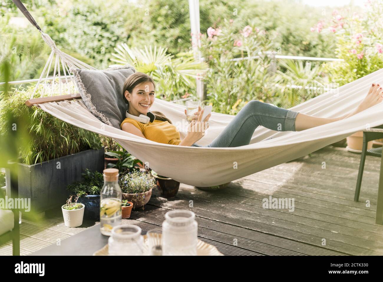 Smiling woman holding drink and smart phone while lying on hammock in yard Stock Photo