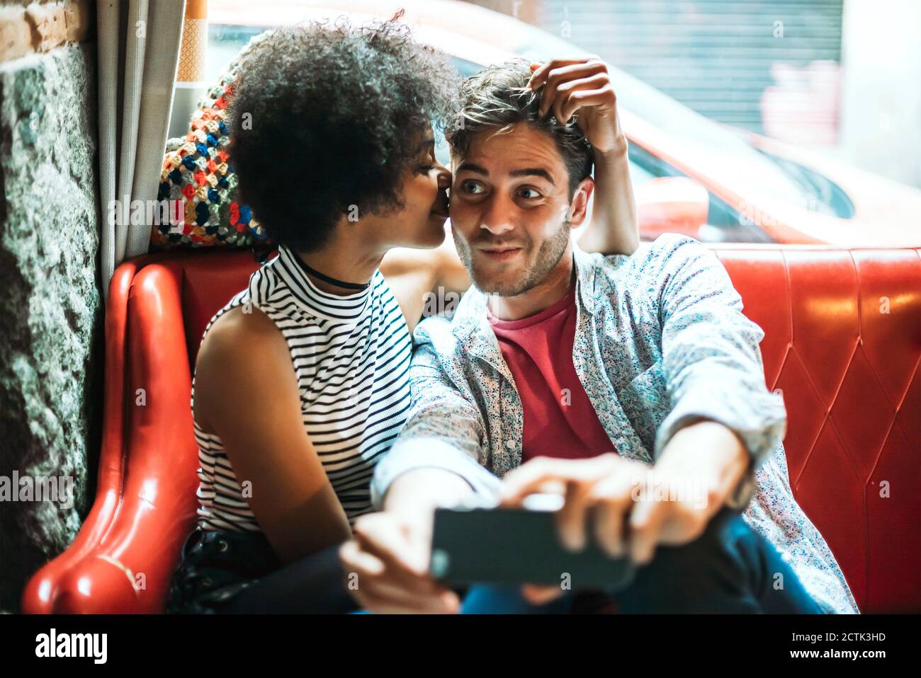 Man taking selfie while girlfriend kissing him while sitting on sofa in cafe Stock Photo