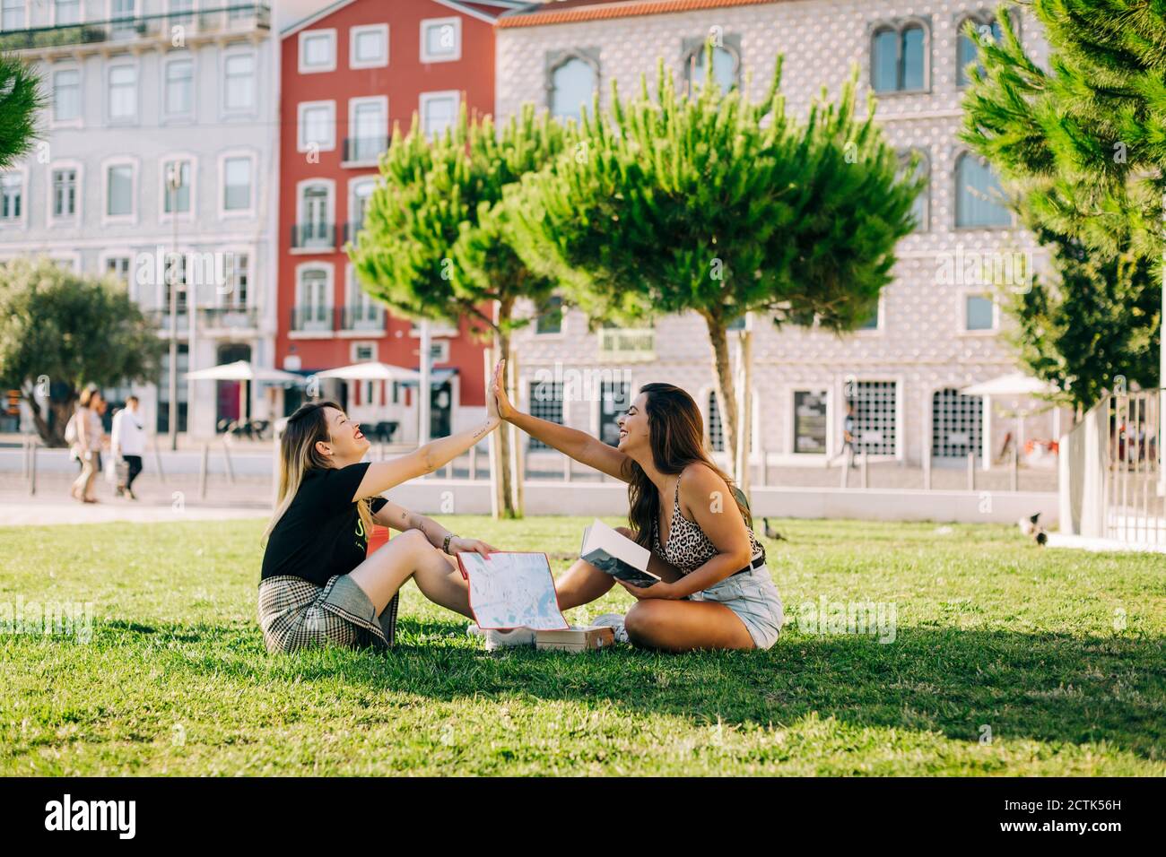 Female tourists giving high-five while sitting on grassy land against building in park Stock Photo